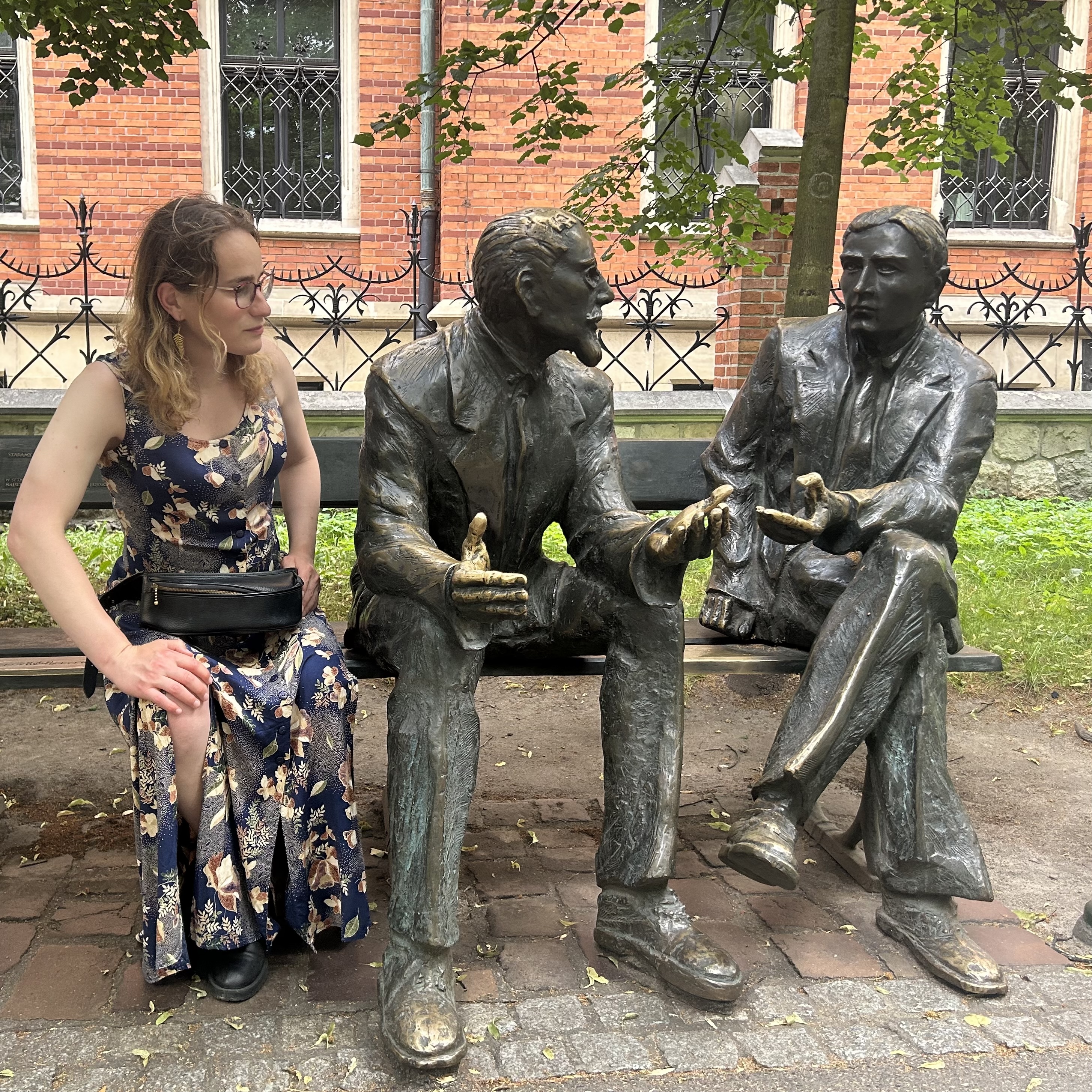 Portrait of a person (Esmeralda S. Whitammer in 2026) on a bench with statues of Stefan Banach and Otto Nikodym in Kraków.